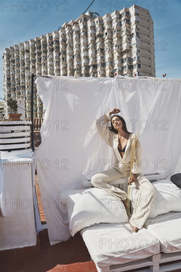 A woman lounges on a rooftop with white linens and cushions, basking in the summer sun The backdrop