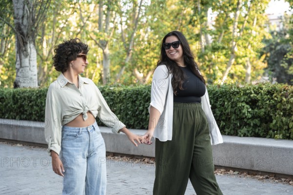 A lesbian couple walks hand-in-hand through a sunny park, enjoying each other's company. The lush