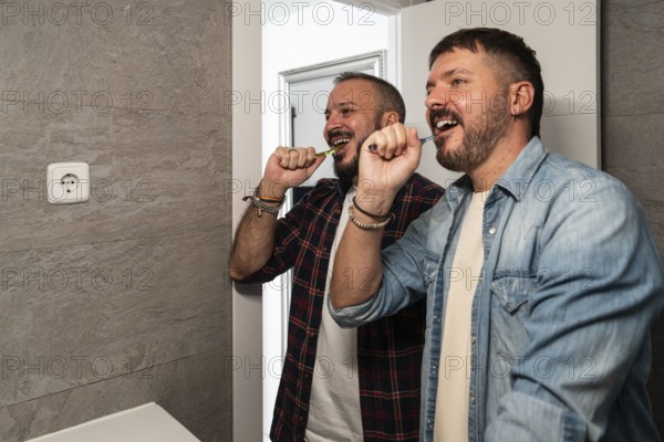 Two gay men in casual shirts joyfully brush their teeth together in a modern bathroom, emphasizing