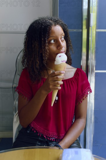 A Mixed-race teenage girl with curly hair, wearing a red blouse, enjoys a strawberry and vanilla