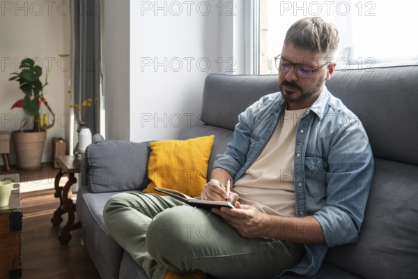A man with glasses sits on a comfortable sofa, writing in a notebook with a pen. Sunlight streams