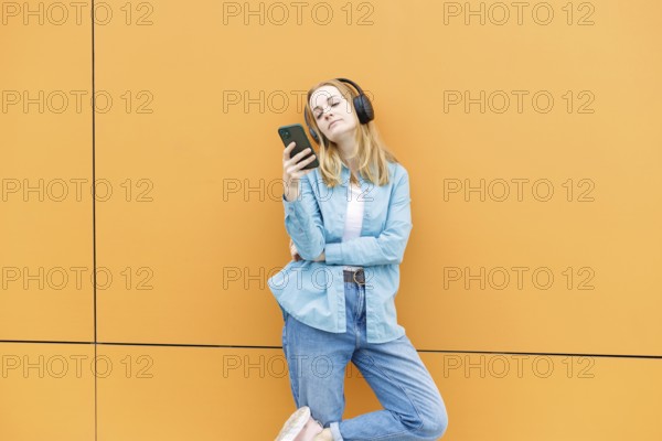 A stylish woman leans against an orange wall in Warsaw, wearing headphones and checking her phone,