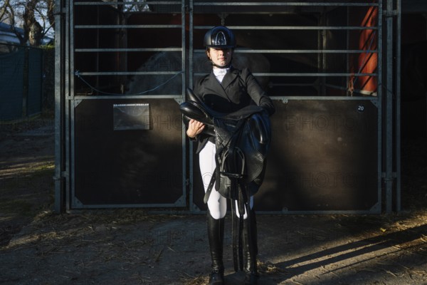 A teenage girl in classical dressage attire confidently holds a saddle. She stands by a stable,