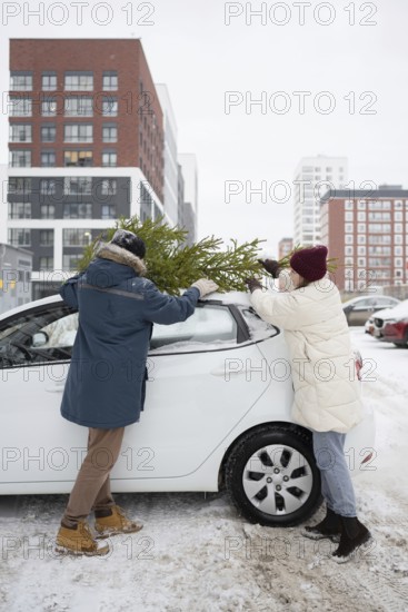 A couple wearing winter clothing secures a Christmas fir onto their car's roof in a snow-covered