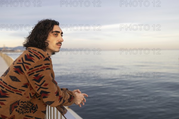 A man with a thoughtful expression leans against a railing, gazing at the tranquil sea The natural