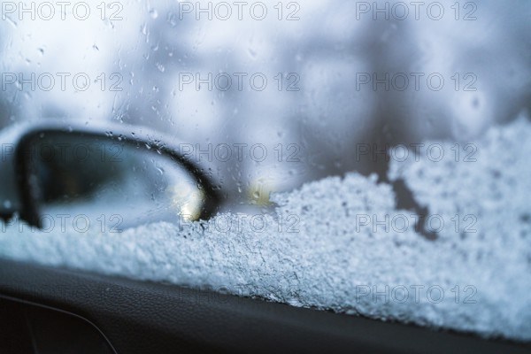 Close-up of a frosted car window with a blurred side mirror visible. The ice crystals and droplets