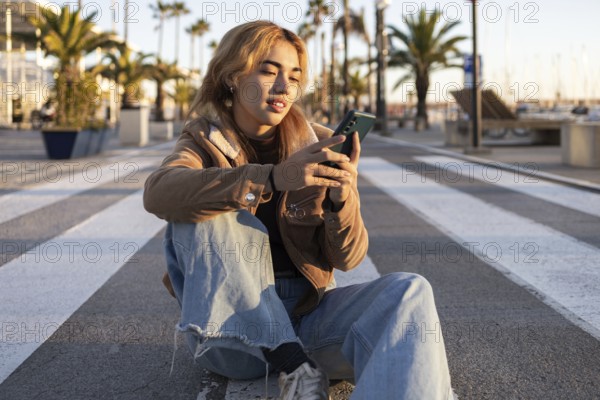 Mixed-race woman sits cross-legged on an outdoor boardwalk, absorbed in her smartphone Palm trees