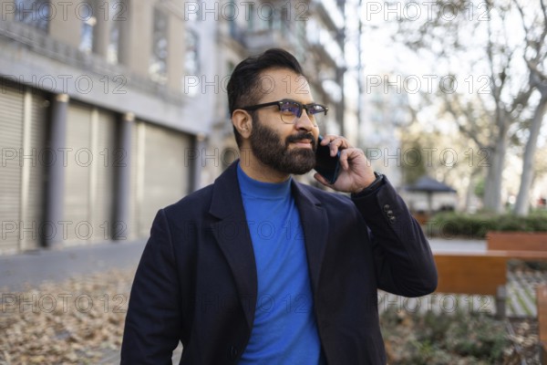 Indian businessman in winter clothing using a mobile phone in an urban park setting, looking away