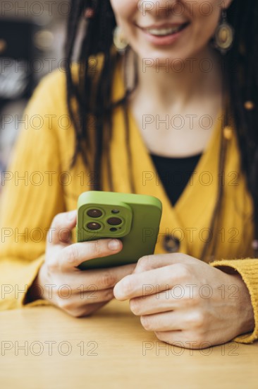 A woman in a mustard yellow sweater holds a smartphone with a green case, smiling The cozy setting