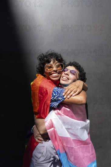 A joyful, multiethnic LGBTQ+ couple embraces warmly, each draped in vibrant pride flags,
