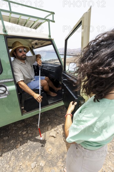 A blind man holds his cane as he steps out of a motorhome, with a woman nearby offering support.