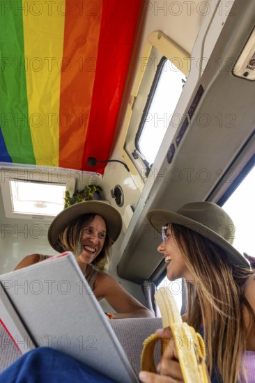 Lesbian couple laughing inside a van decorated with a rainbow flag. One holds a book while the