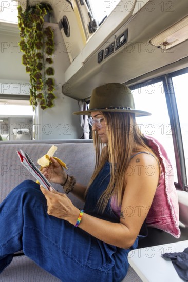 A lesbian woman in a hat and overalls enjoys a banana while reading a tablet inside a cozy camper
