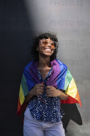 A smiling LGTBQ+ person wearing sunglasses and a patterned shirt, joyfully wrapped in a rainbow