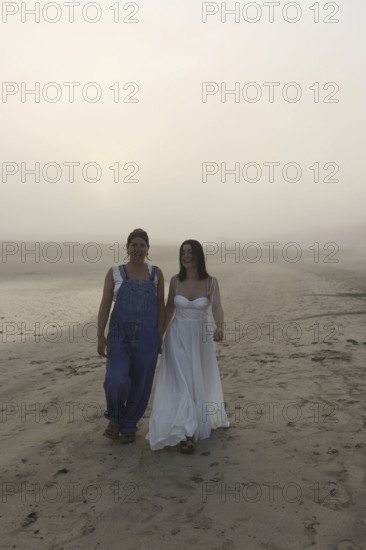Two women walk along a foggy beach, one in overalls and the other in a flowing white dress The
