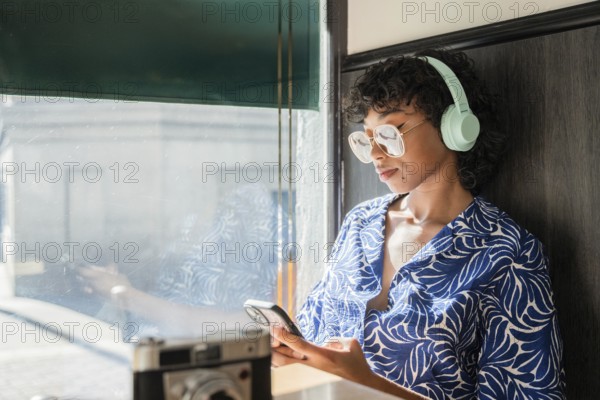 A stylish young woman with curly hair and glasses wearing headphones, leans against a window while