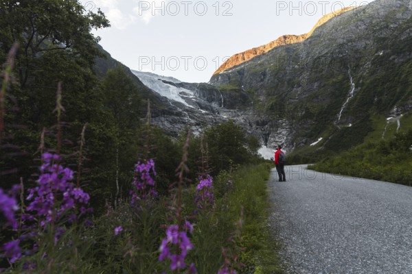 A traveler in a red jacket stands on a scenic mountain path in Norway, surrounded by vibrant