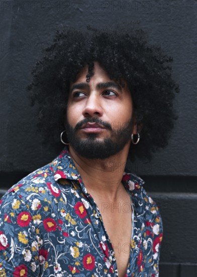 Portrait of a stylish man with curly hair wearing hoop earrings and a vibrant floral shirt. The