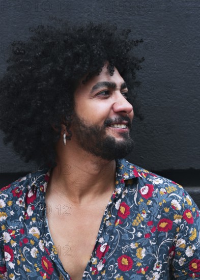 A man with curly hair and a beard smiles while wearing a vibrant floral shirt against a gray