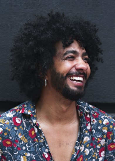 A smiling man with curly hair and a floral shirt exudes happiness. Their joyful expression and