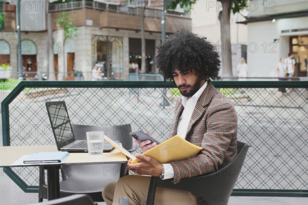 A Mixed-race man with curly hair is engaged in remote work using a laptop and smartphone at an