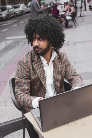 A Mixed-race man with curly hair, dressed in a stylish plaid jacket and white shirt, is focused on