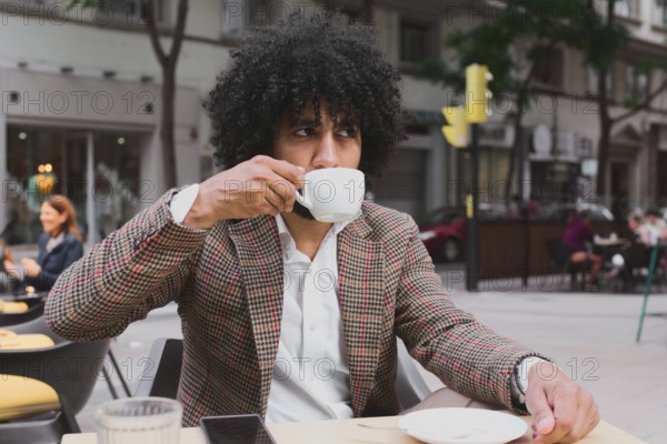 A Mixed-race man with curly hair teleworks from a street cafe, sipping coffee, dressed in a stylish