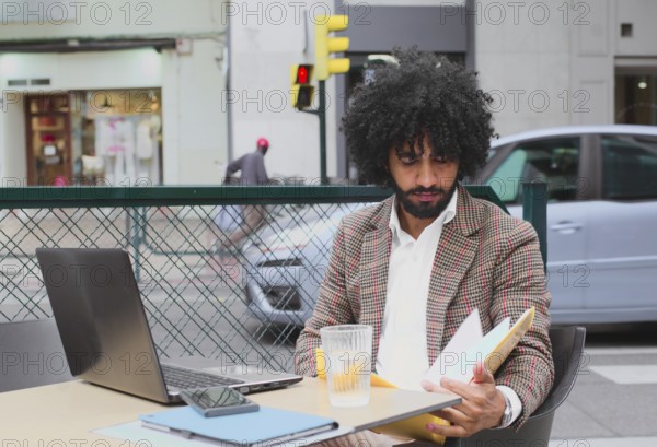 A young Mixed-race man with curly hair working remotely at a street-side cafe, carefully reviewing
