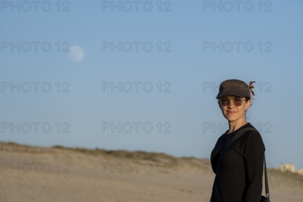 A smiling woman dressed in casual clothes and sunglasses stands on a sandy beach looking at camera