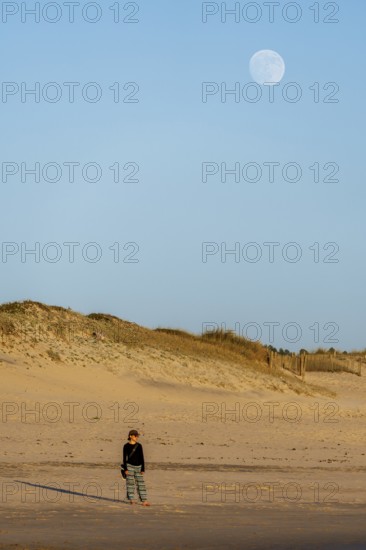 A peaceful scene of a woman standing on the sandy beach at twilight, with a large moon rising above