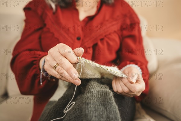 A woman in a red blouse sits comfortably on a couch, skillfully knitting a wool sweater. The