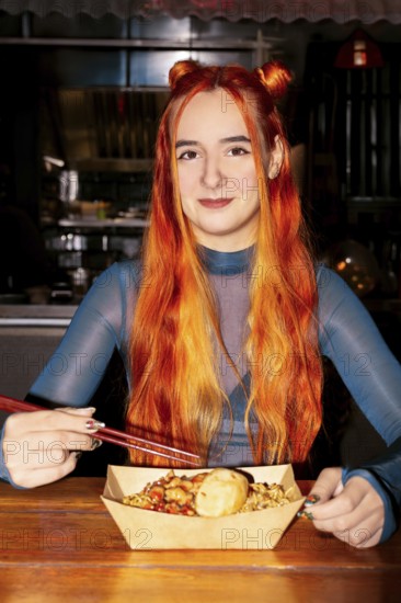 A woman with vibrant red hair sits at an Asian food market, savoring a meal with chopsticks The