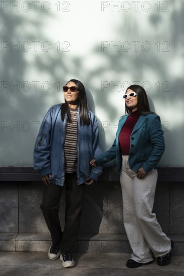 A stylish Latin lesbian couple enjoys a sunny day, dressed in casual fashion with jackets and