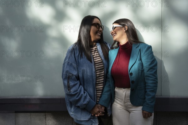 A Latin lesbian couple stands closely, smiling at each other with affection. They hold hands,