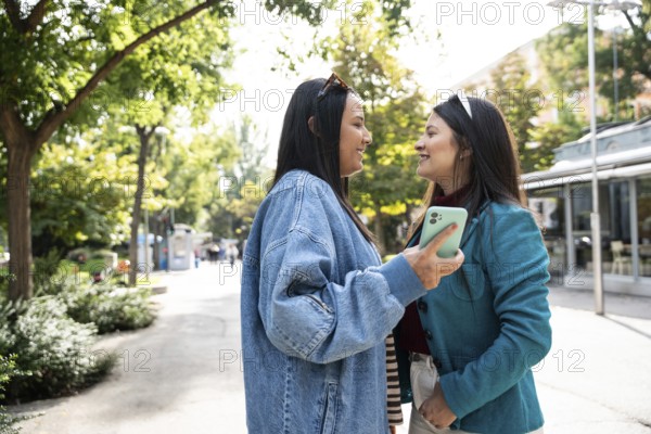A cheerful Latin lesbian couple stands close outdoors, sharing a joyful moment. They smile warmly