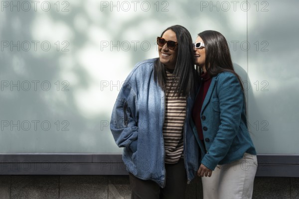 A Latin lesbian couple stands close, basking in the sun. One wears a denim jacket, the other a teal