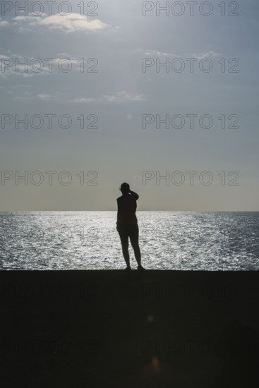 Silhouette of a person standing by the sea during sunset. The calm water and warm light create a