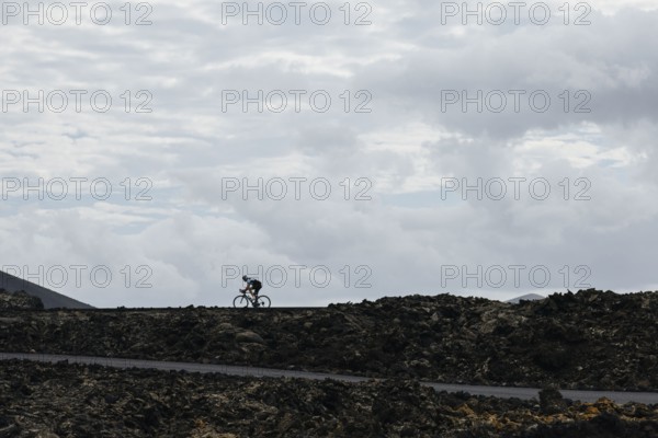 A lone cyclist riding on a volcanic road under a cloudy sky. The contrast between the dark terrain