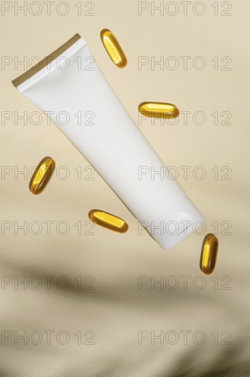 Top view of a blank white cosmetic tube floats amidst golden oil capsules, all set against a soft
