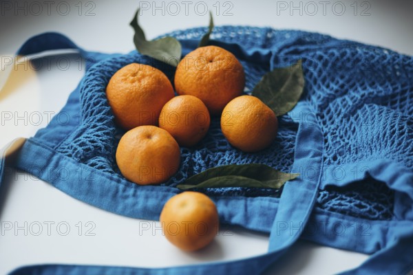 Fresh tangerines with green leaves arranged in a vibrant blue string bag, set against a white table