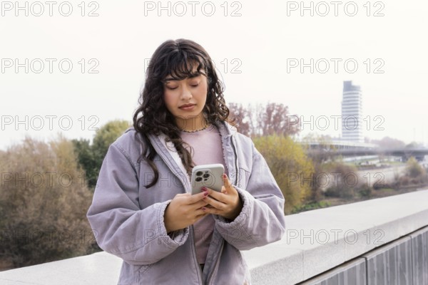 Young Latin woman in a casual jacket checking her phone outdoors, standing on a bridge with trees