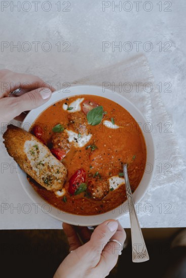 From above cropped unrecognizable person holding a bowl of tomato soup with meatballs, bread, and