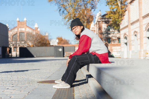 A young Latina woman in a red shirt and black pants sits on a concrete bench in an urban area It's