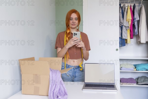 A young woman organizes clothes for online resale, using a laptop and smartphone. A box is ready