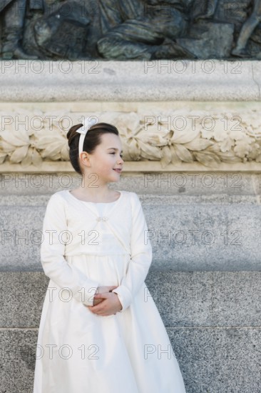 A young girl stands in a beautiful First Communion dress, framed by an ornate stone wall with