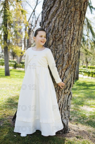 A young girl stands by a tree, smiling in a beautiful white First Communion dress. Sunlight filters