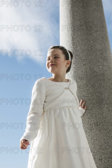 A young girl wearing a white First Communion dress stands outdoors against a stone column. The