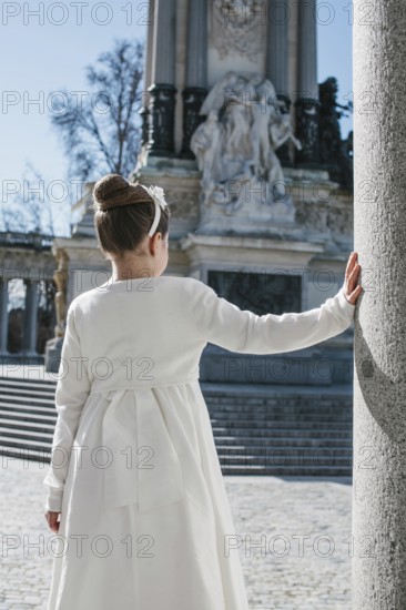 A young girl wearing a white First Communion dress stands beside a stone column, with a historic