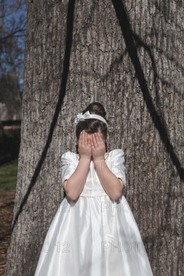 A young girl stands in front of a tree, covering her face with her hands. She is wearing an elegant
