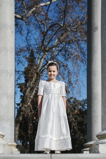 A young girl stands in a white First Communion dress, framed by stone columns and bare trees, with